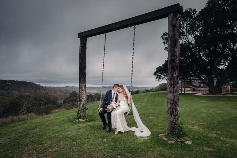 Chelsea the bride and Brent the groom sit closely together on a large wooden swing at Preston Peak Winery Functions Centre, with a scenic view of rolling hills and trees under a cloudy sky.