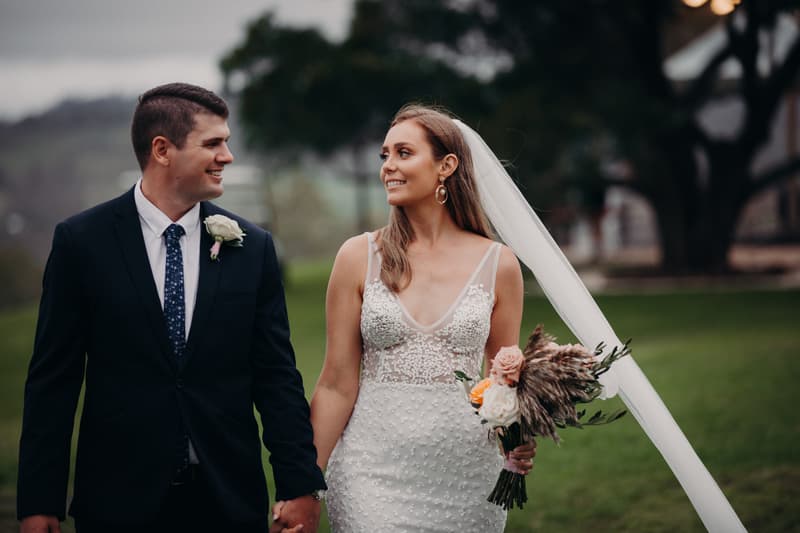 Chelsea and Brent walk hand in hand outdoors at Preston Peak Winery Functions Centre, with Chelsea holding a bouquet and wearing a white wedding dress and veil, and Brent in a dark suit with a boutonniere.