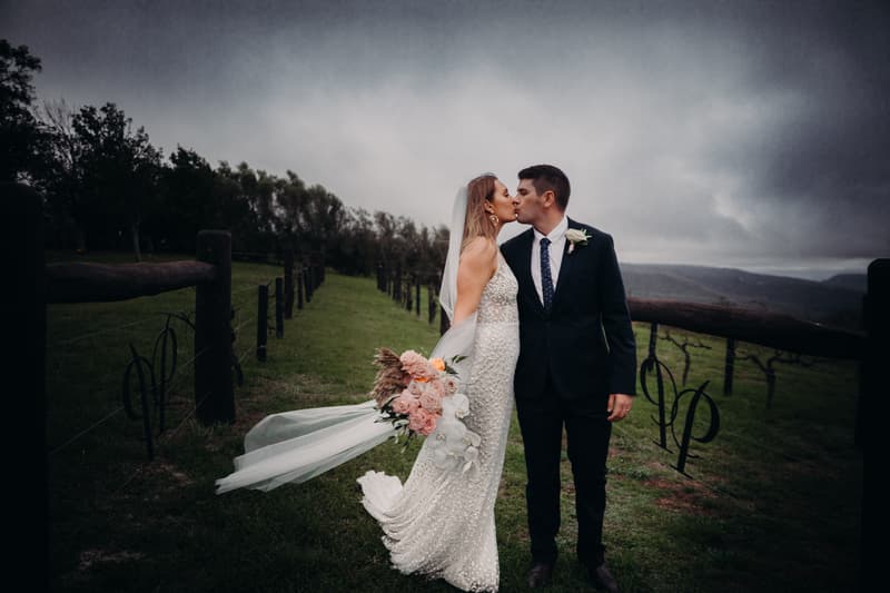 Chelsea and Brent kiss during their couple portraits session at Preston Peak Winery Functions Centre, standing on a grassy path between wooden fences with vineyard hills in the background under a cloudy sky. Chelsea wears a white wedding gown and veil, holding a bouquet of flowers, while Brent is dressed in a dark suit with a boutonniere.
