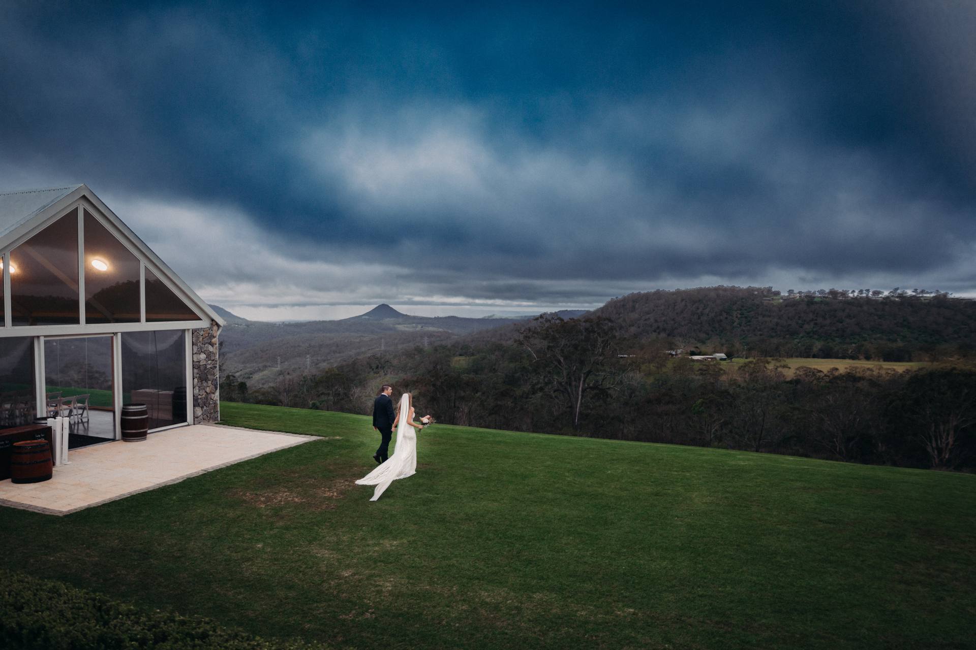 The bride Chelsea and groom Brent walk together on a grassy area outside the Preston Peak Winery Functions Centre with a scenic mountainous landscape under a cloudy sky.