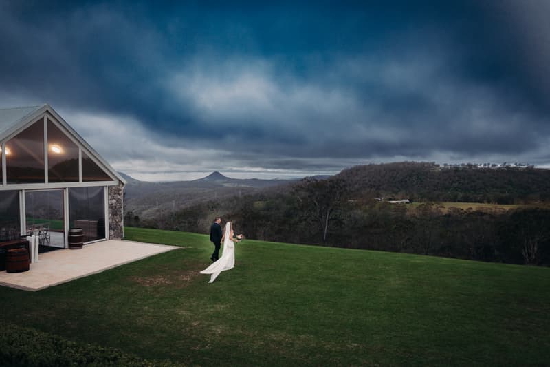 The bride Chelsea and groom Brent walk together on a grassy area outside the Preston Peak Winery Functions Centre with a scenic mountainous landscape under a cloudy sky.