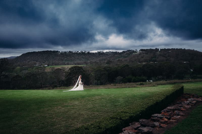 The bride Chelsea stands alone on a large grassy area at Preston Peak Winery Functions Centre, wearing a long white wedding gown and veil, holding a bouquet, with a backdrop of hills and a cloudy sky.