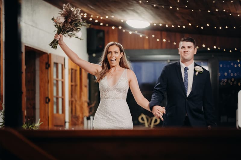 Chelsea the bride and Brent the groom hold hands as Chelsea raises her bouquet inside Preston Peak Winery Functions Centre, with string lights overhead and wooden doors in the background.