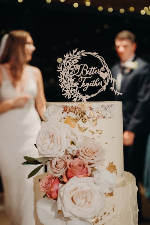 Wedding cake decorated with white and pink roses and gold leaf, topped with a 'Better Together' sign, with the bride and groom blurred in the background at Preston Peak Winery Functions Centre.
