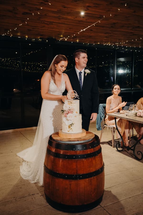 Bride Chelsea and groom Brent cut their wedding cake on a wooden barrel at Preston Peak Winery Functions Centre, while a bridesmaid seated at a table claps.