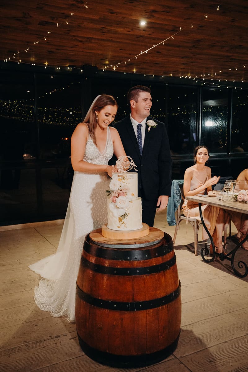 Bride Chelsea and groom Brent cut their wedding cake on a wooden barrel, while a bridesmaid seated at a table claps. Brisbane wedding photographers by She Said Yes.