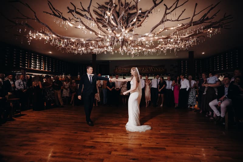 Bride Chelsea and groom Brent dance together on the wooden floor at Preston Peak Winery Functions Centre, surrounded by guests watching.