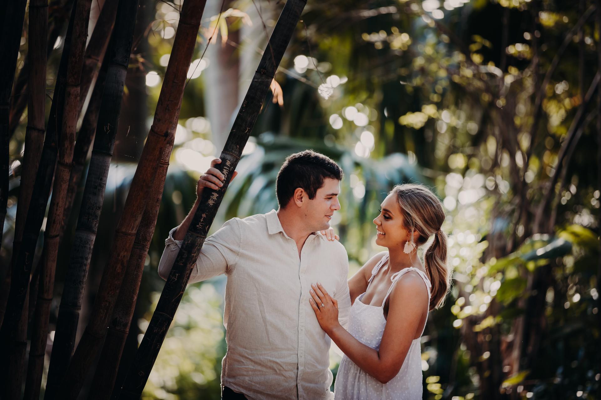 The couple stands close together in Brisbane Botanic Gardens, surrounded by lush greenery, with soft sunlight filtering through the trees.