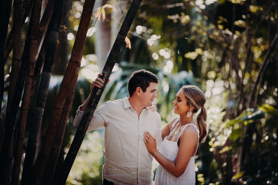 The couple stands close together in Brisbane Botanic Gardens, surrounded by lush greenery, with soft sunlight filtering through the trees.
