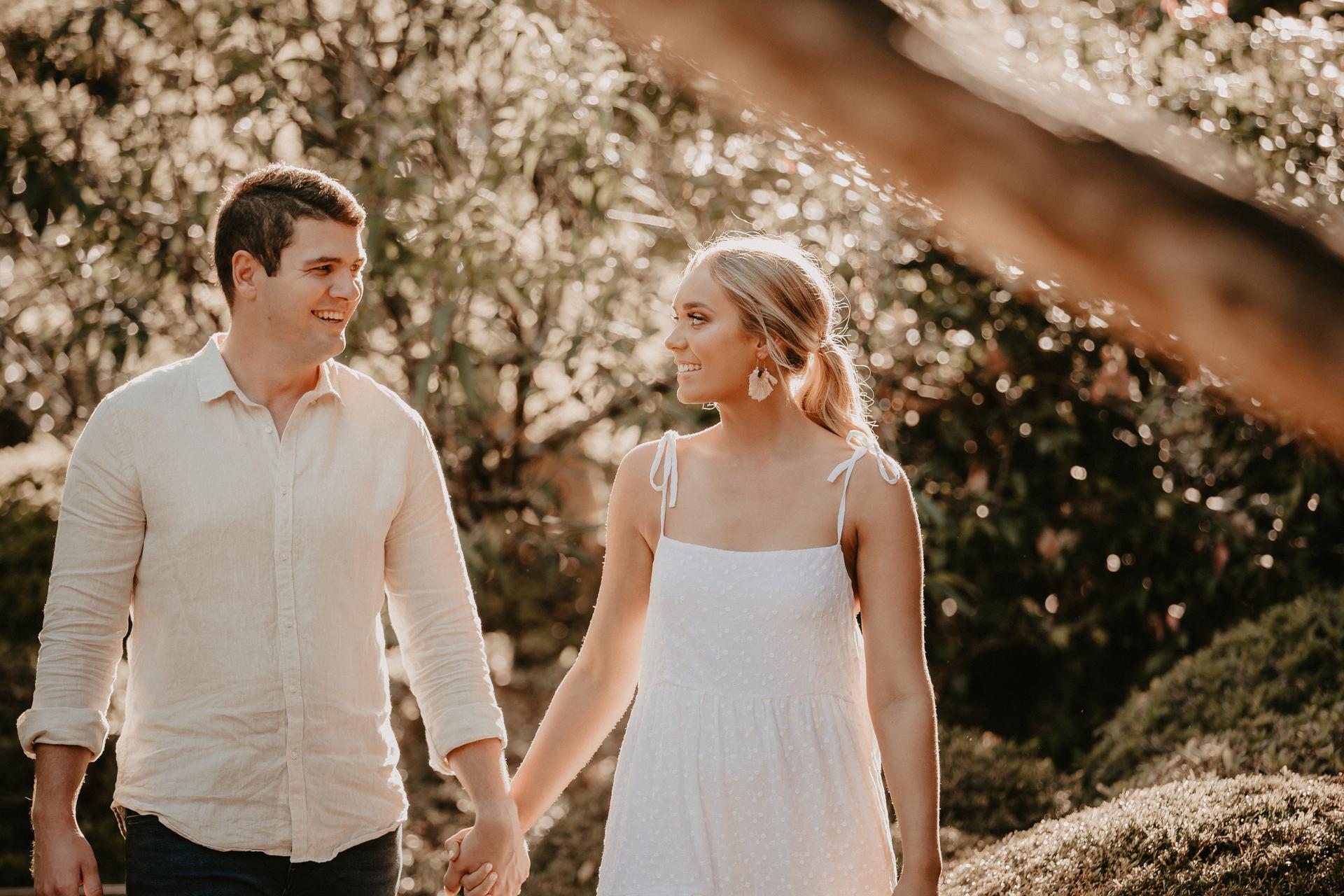 The couple walks hand in hand through the Brisbane Botanic Gardens, smiling at each other under the dappled sunlight.