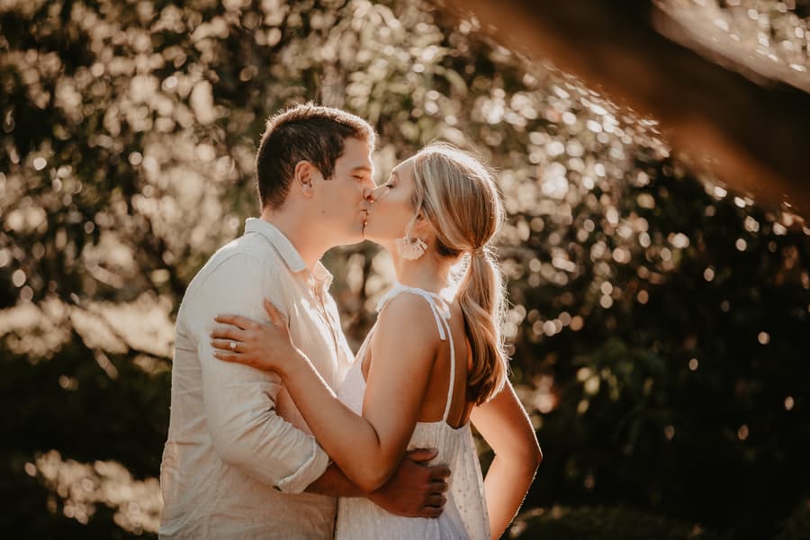 The couple shares a kiss in the Brisbane Botanic Gardens, surrounded by lush foliage and warm sunlight.