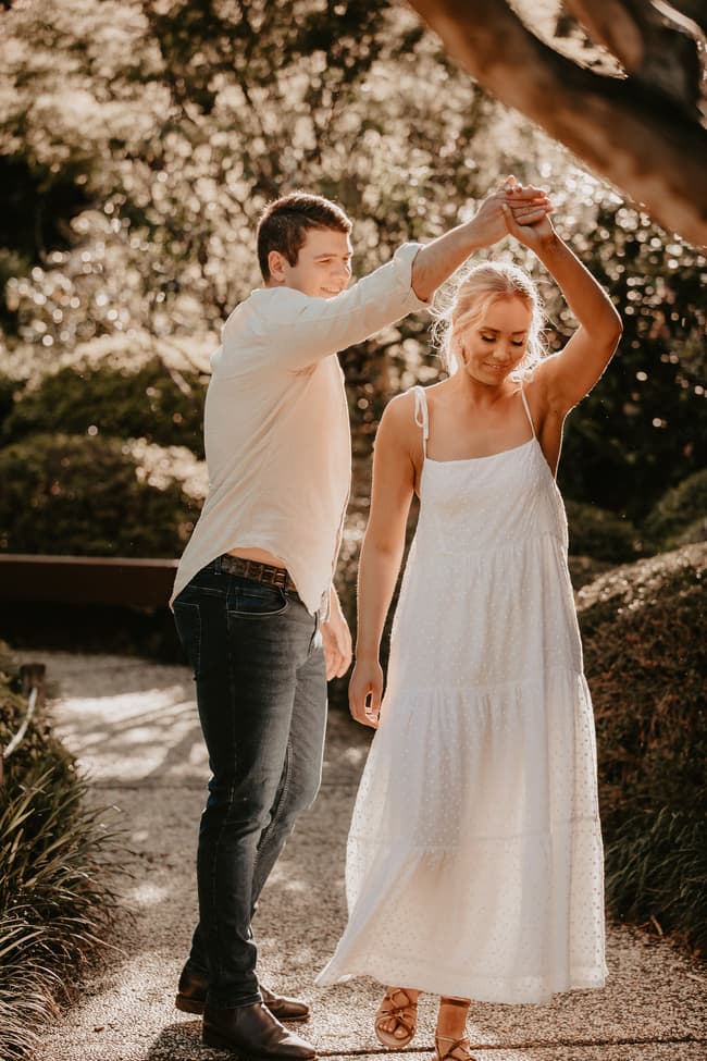 The couple dances playfully on a garden path in the Brisbane Botanic Gardens, with sunlight filtering through the trees.