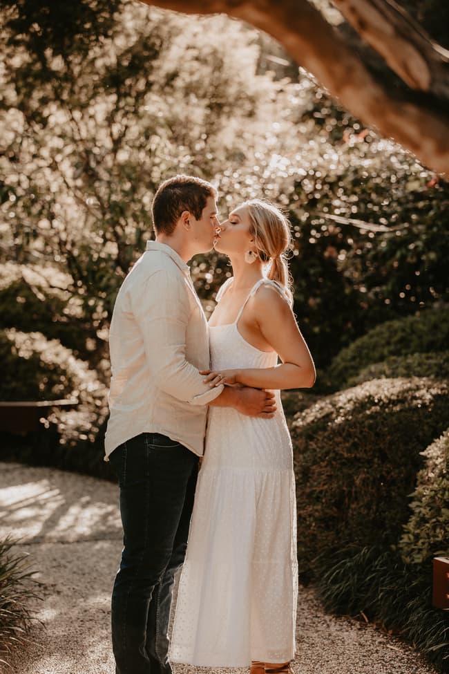 The couple shares a tender kiss on a garden path in the Brisbane Botanic Gardens, surrounded by greenery.