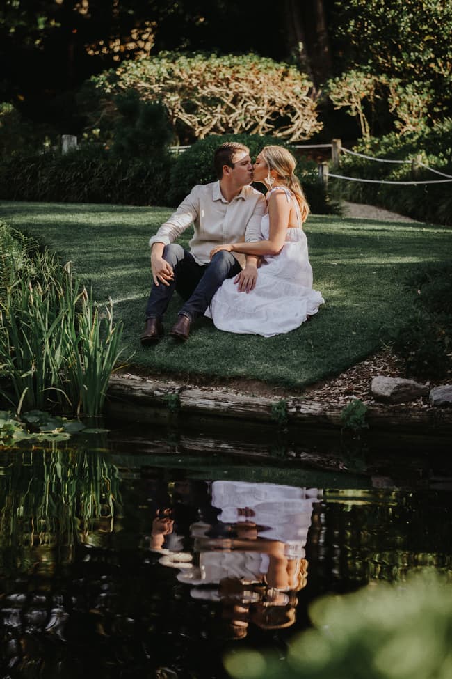 The couple sits by a pond in the Brisbane Botanic Gardens, sharing a kiss with their reflection visible in the water.