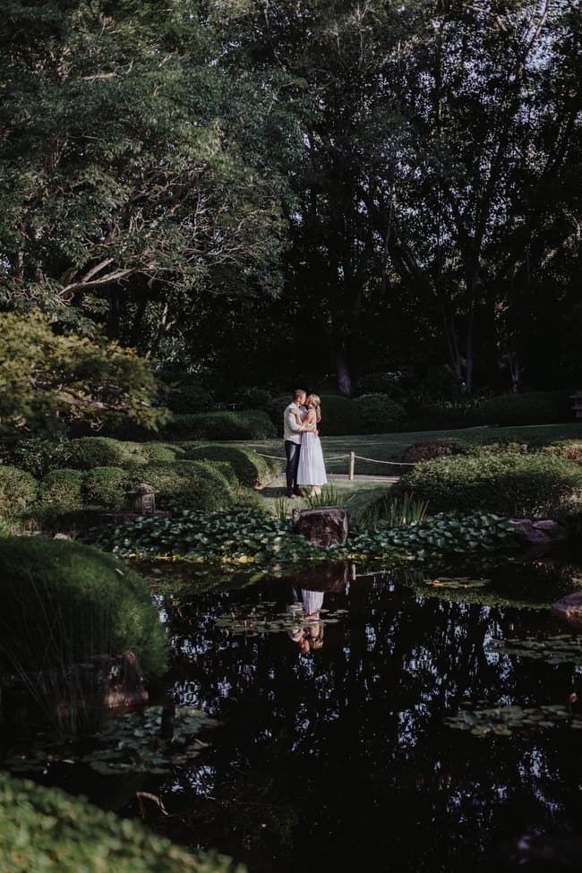 The couple stands embraced by a pond in the Brisbane Botanic Gardens, surrounded by lush greenery.