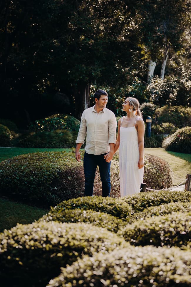 The couple walks hand in hand through the Brisbane Botanic Gardens, surrounded by manicured bushes and trees.