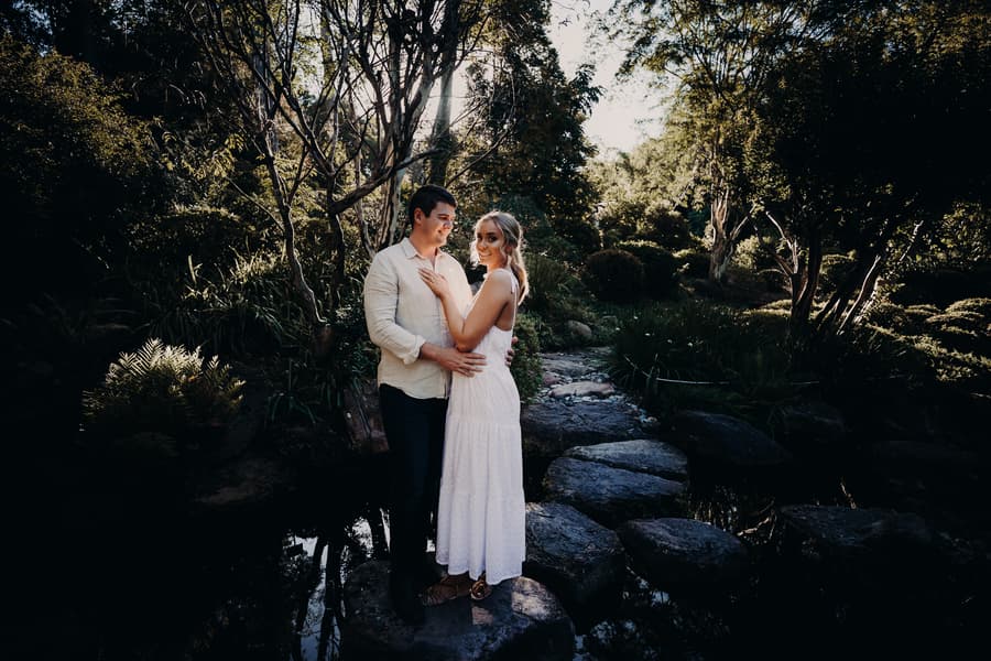 The couple stands on rocks by a pond, embraced under the dappled sunlight at Brisbane Botanic Gardens.