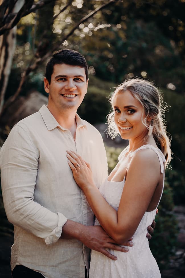 The couple smiles warmly at the camera, standing close together in a sunlit garden setting.