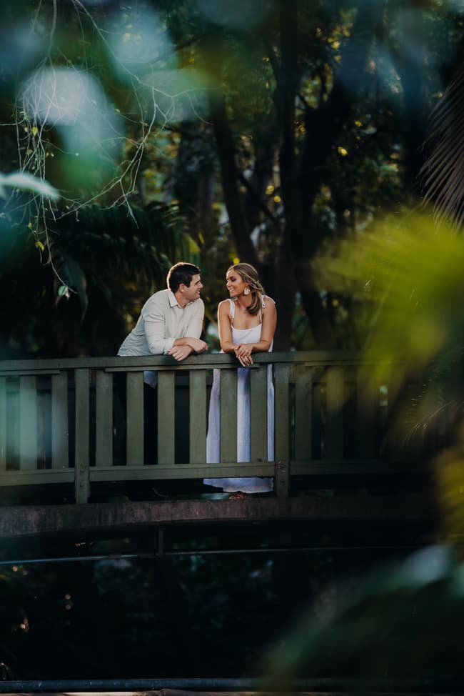 The couple leans on a wooden bridge, gazing into each other's eyes amidst lush foliage.