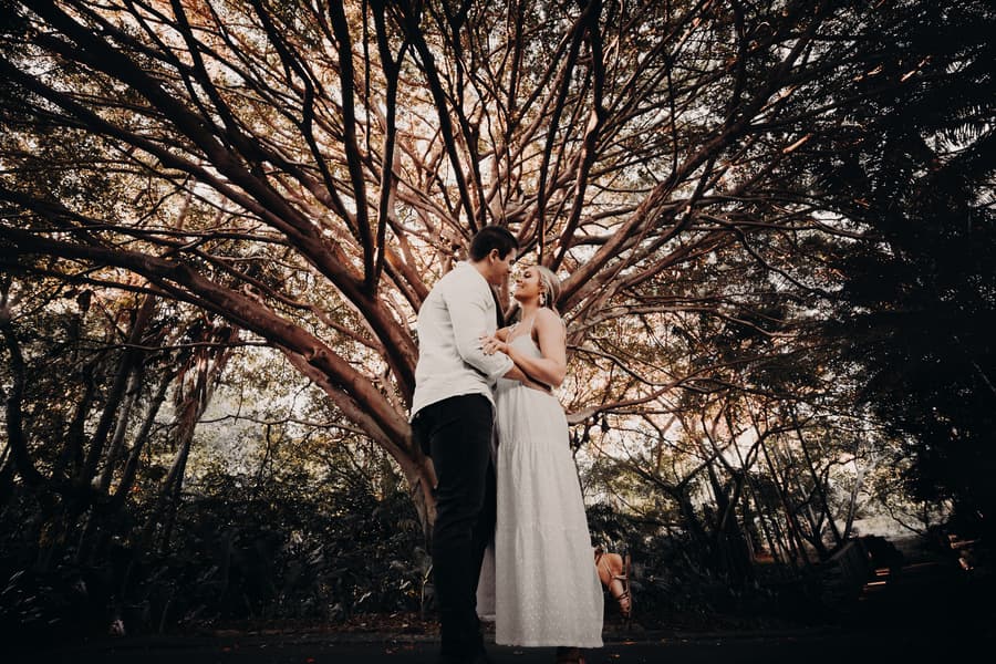 The couple stands under a large tree, embraced and looking at each other lovingly.
