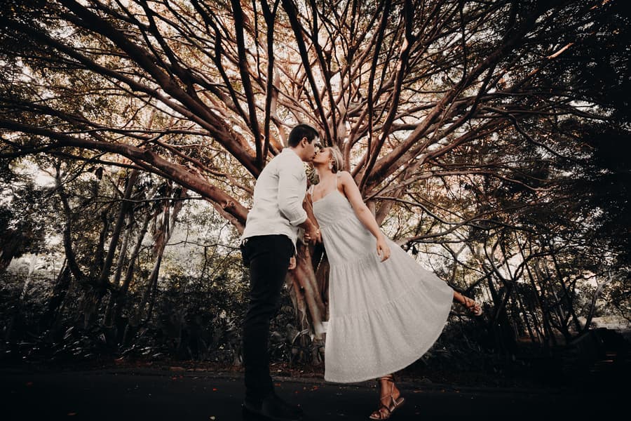 The couple shares a playful kiss under a sprawling tree, one person lifting a foot playfully.