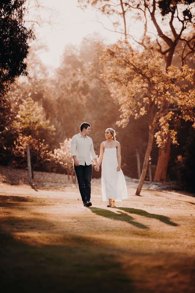 The couple walks hand in hand on a sunlit path, surrounded by trees and soft light.
