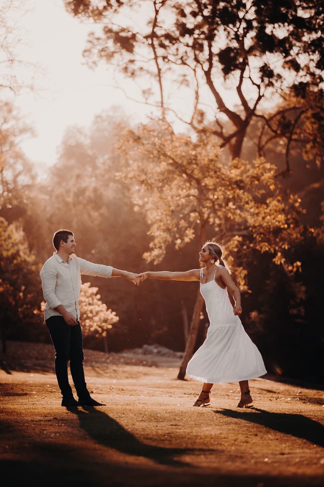 The couple holds hands, one reaching out playfully, surrounded by trees and warm sunlight.