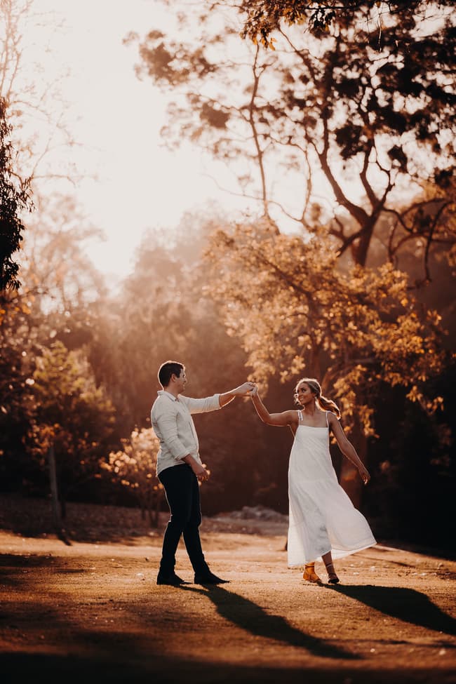 The couple dances gracefully on a sunlit lawn, surrounded by trees and soft light.