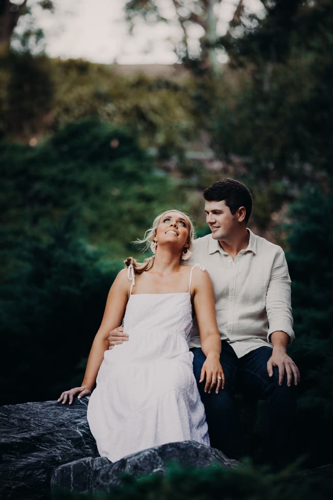 The couple sits on a rock, with one looking up and the other gazing at them, surrounded by greenery.