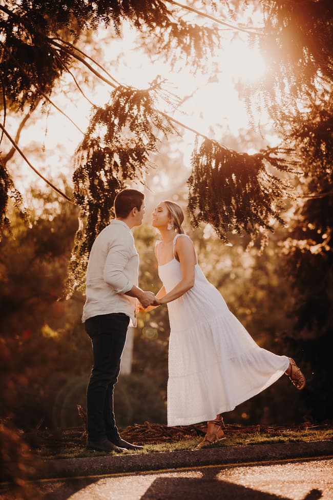 The couple shares a playful moment, holding hands and leaning in for a kiss under sunlit trees.