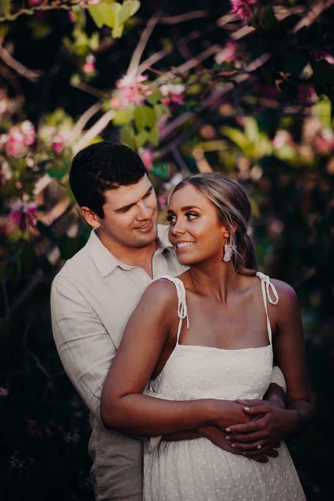 The couple embraces among flowering branches, exchanging warm smiles in the garden setting.