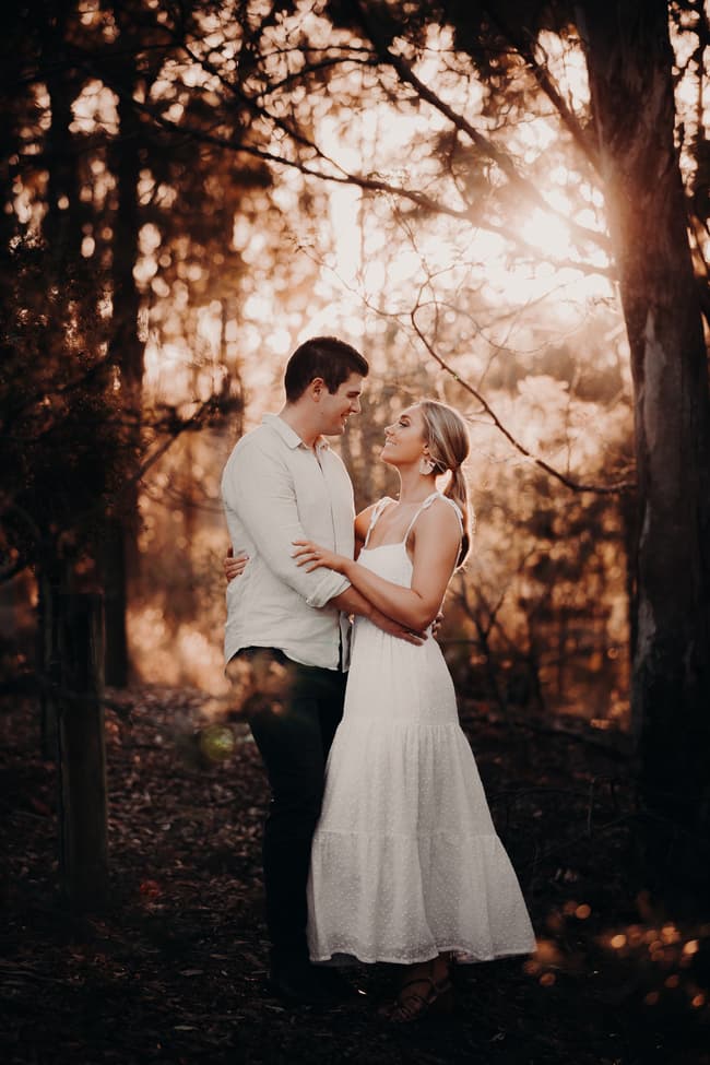 The couple stands in a wooded area, gazing at each other with affection as sunlight filters through.