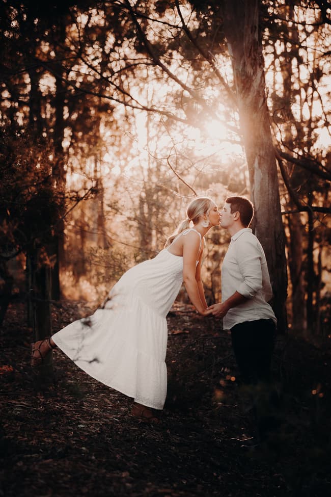 The couple shares a kiss, with one playfully leaning forward, surrounded by trees and warm light.