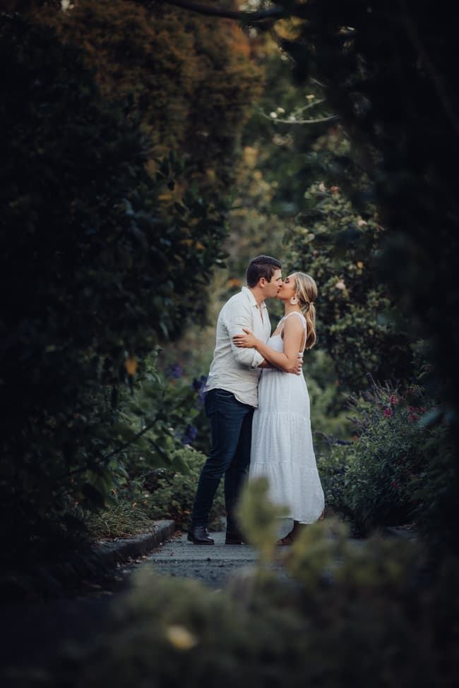 The couple shares a kiss on a garden path, framed by lush foliage and vibrant flowers.