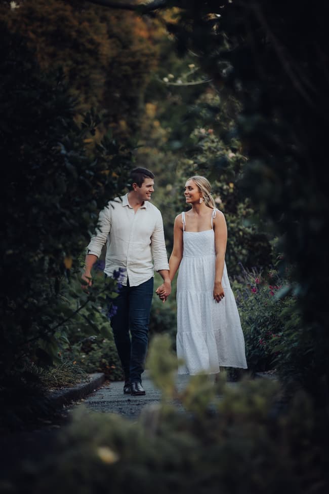The couple walks hand in hand along a garden path, surrounded by greenery and flowers.