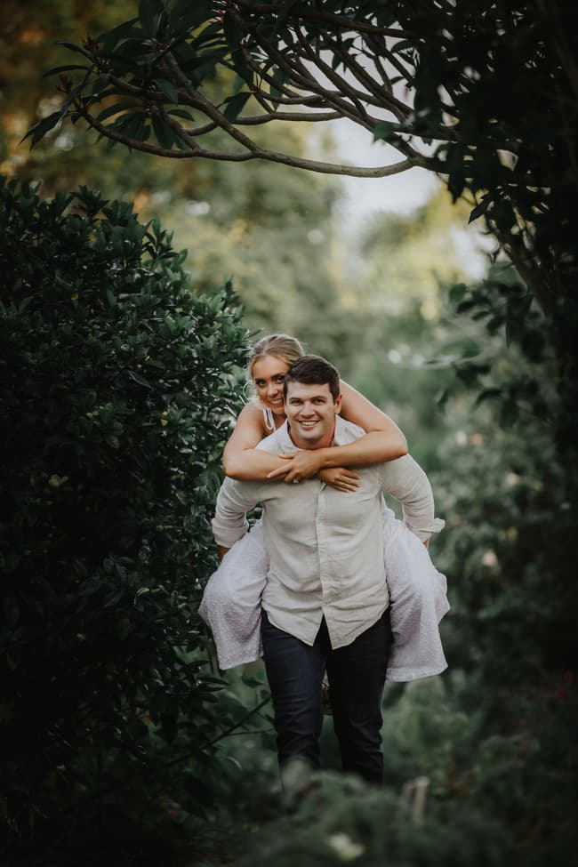 The couple shares a playful piggyback ride, smiling as they move through a lush garden setting.