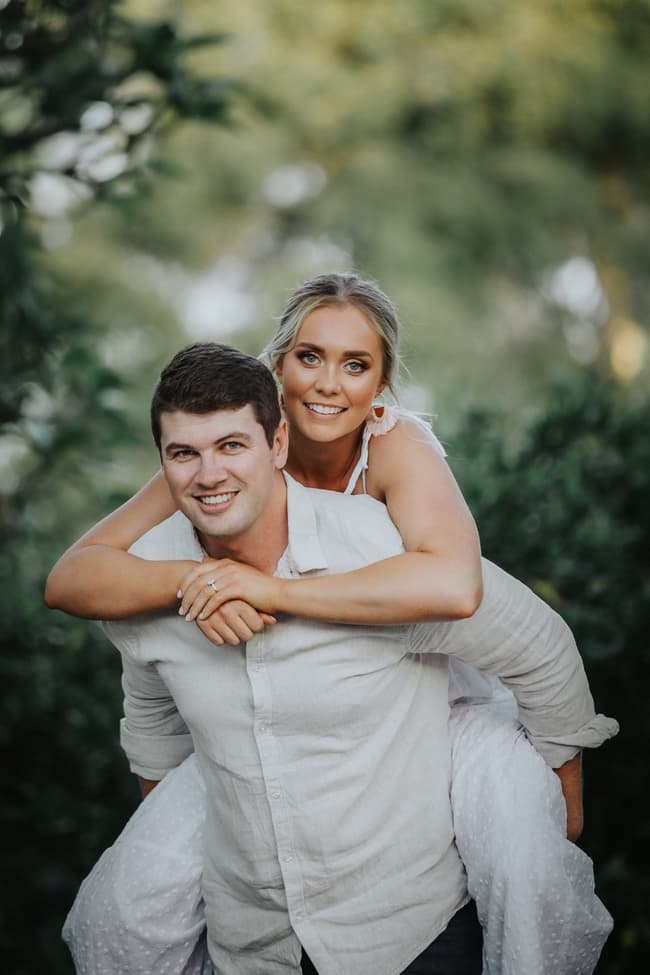 The couple in a playful piggyback pose, smiling warmly, surrounded by lush greenery at Brisbane Botanic Gardens.