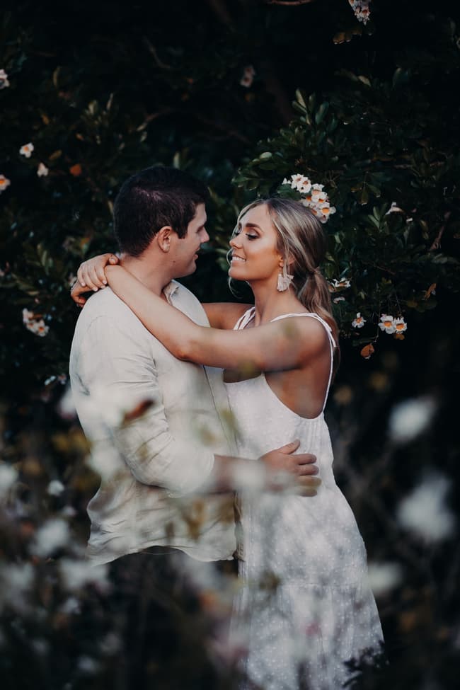 The couple embracing tenderly, gazing into each other's eyes, with flowering bushes in the background.
