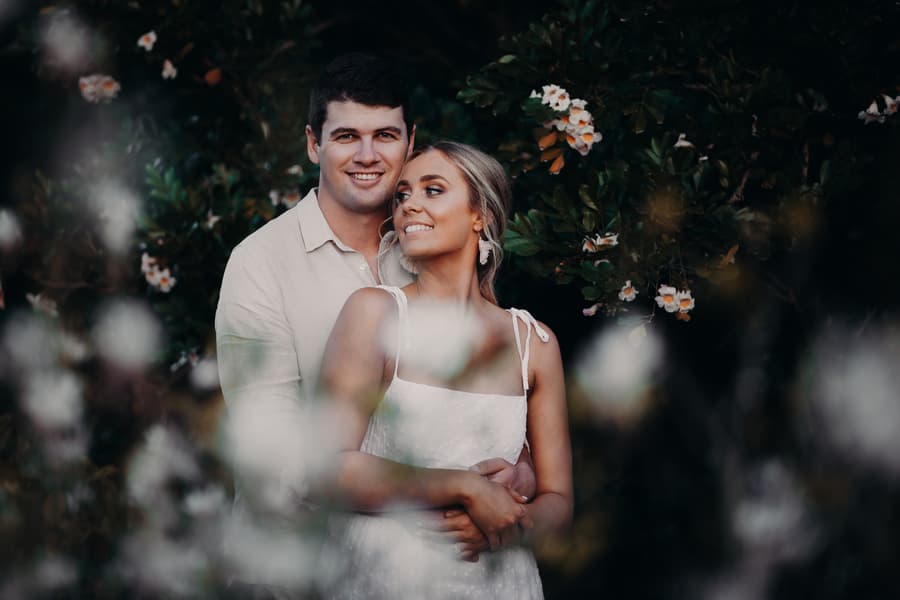 The couple standing closely, smiling softly, surrounded by blurred flowers and greenery.
