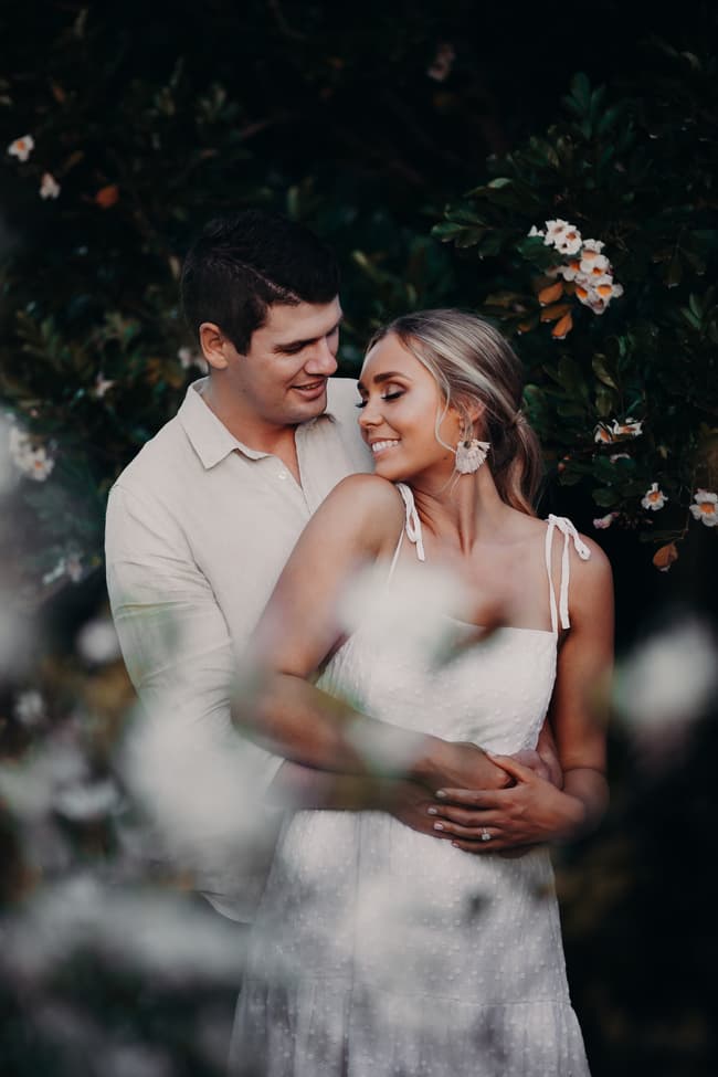 The couple sharing a loving moment, embraced with smiles, amidst lush foliage and flowers.