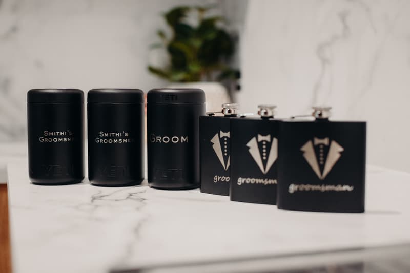 Black YETI drink holders labeled 'Smithi's Groomsmen' and 'Groom' alongside black flasks with tuxedo designs and the word 'groomsman' on a marble surface.