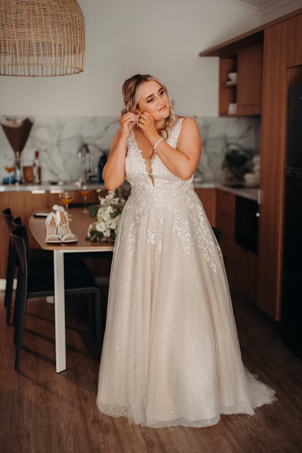 The bride Chloe adjusts her earring in a kitchen area at Sandstone Point Hotel while wearing her wedding gown. A pair of white bridal shoes and a bouquet are on the table behind her.