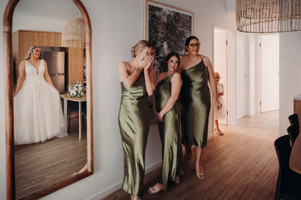 The bride Chloe stands in a room near a mirror wearing her wedding gown, while three bridesmaids in matching green dresses react emotionally and smile nearby inside a modern indoor space. Wedding photographer Brisbane by She Said Yes.