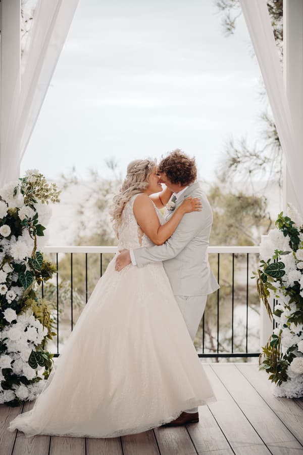 The bride Chloe and groom Brodie embrace and kiss at the altar during their wedding ceremony at Sandstone Point Hotel — Pavilion, framed by white floral arrangements and sheer drapes.