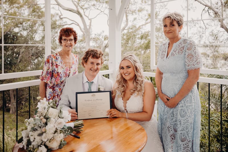 Brodie and Chloe sit at a wooden table holding their marriage certificate at Sandstone Point Hotel — Pavilion, flanked by two older women standing behind them, with a bridal bouquet on the table.