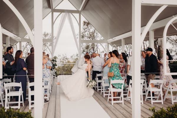 The bride Chloe and groom Brodie share a kiss at the end of the ceremony aisle at Sandstone Point Hotel — Pavilion, surrounded by standing guests on both sides under a white pavilion structure.