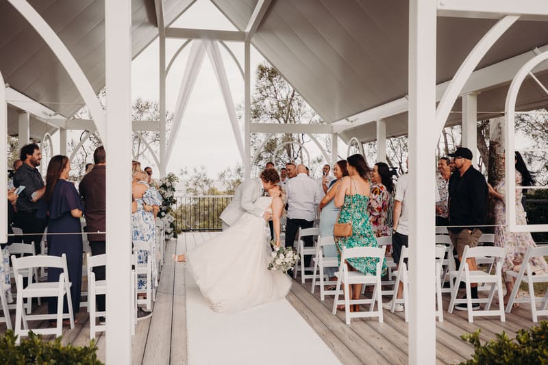 The bride Chloe and groom Brodie share a kiss at the end of the ceremony aisle, surrounded by standing guests on both sides under a white pavilion structure. Good wedding vows by She Said Yes.