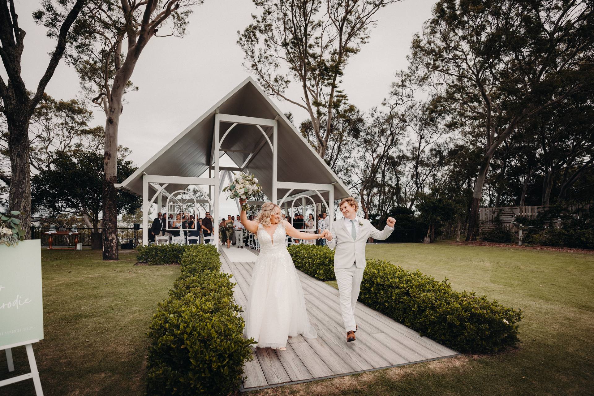 Bride Chloe and groom Brodie walk hand in hand down the aisle at Sandstone Point Hotel — Pavilion after the ceremony, with guests seated under the pavilion in the background.