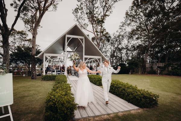 Bride Chloe and groom Brodie walk hand in hand down the aisle at Sandstone Point Hotel — Pavilion after the ceremony, with guests seated under the pavilion in the background.