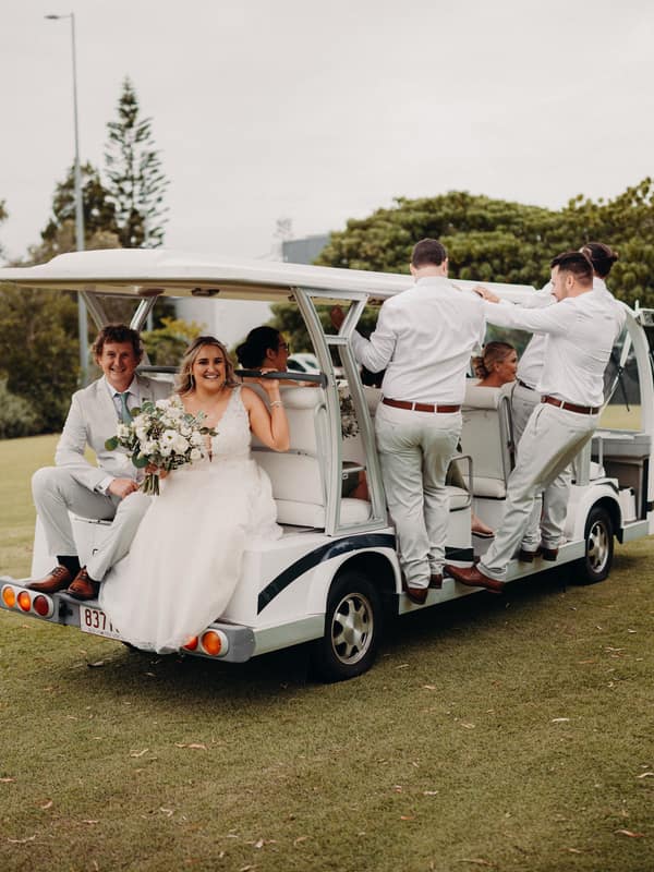 The bride Chloe and groom Brodie sit at the back of a white golf cart at Sandstone Point Hotel, accompanied by wedding party members dressed in light-colored attire, some standing on the side steps of the cart.